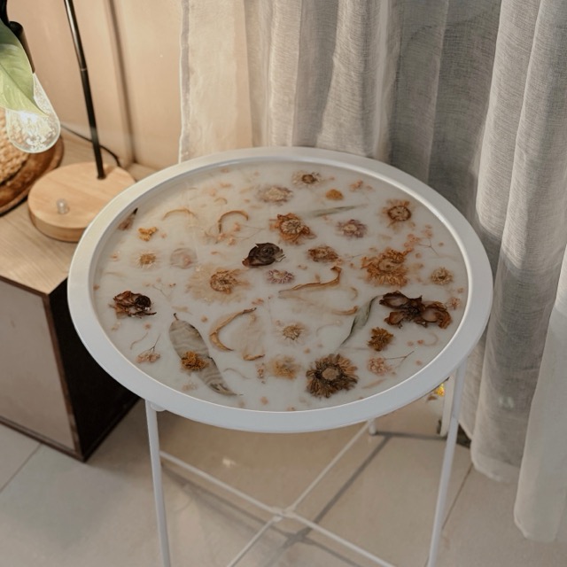White round side table with a resin top holding preserved flowers, beside a sheer curtain