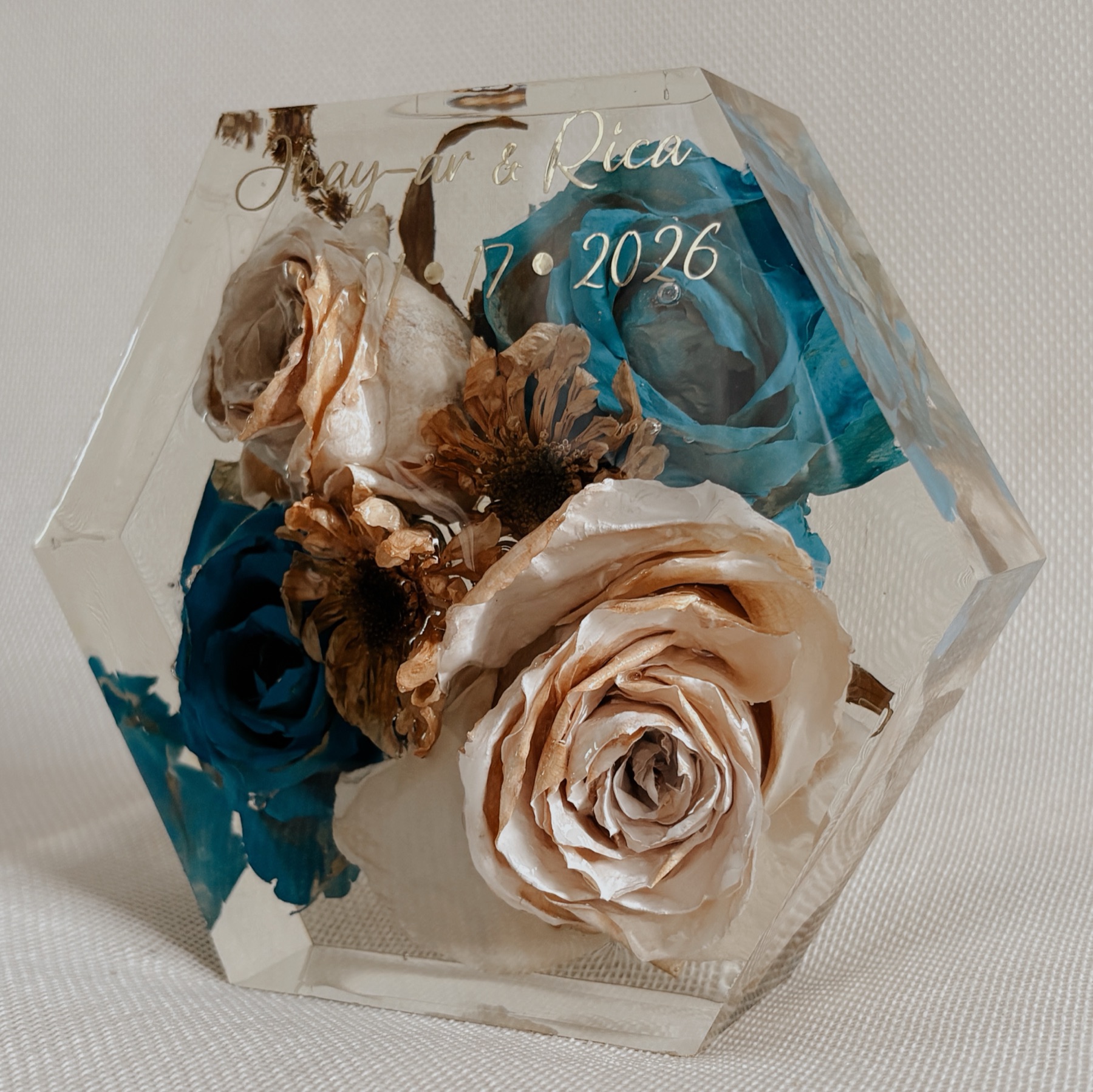 A hexagonal resin block holding preserved cream and teal roses, engraved with a couple's names and wedding date